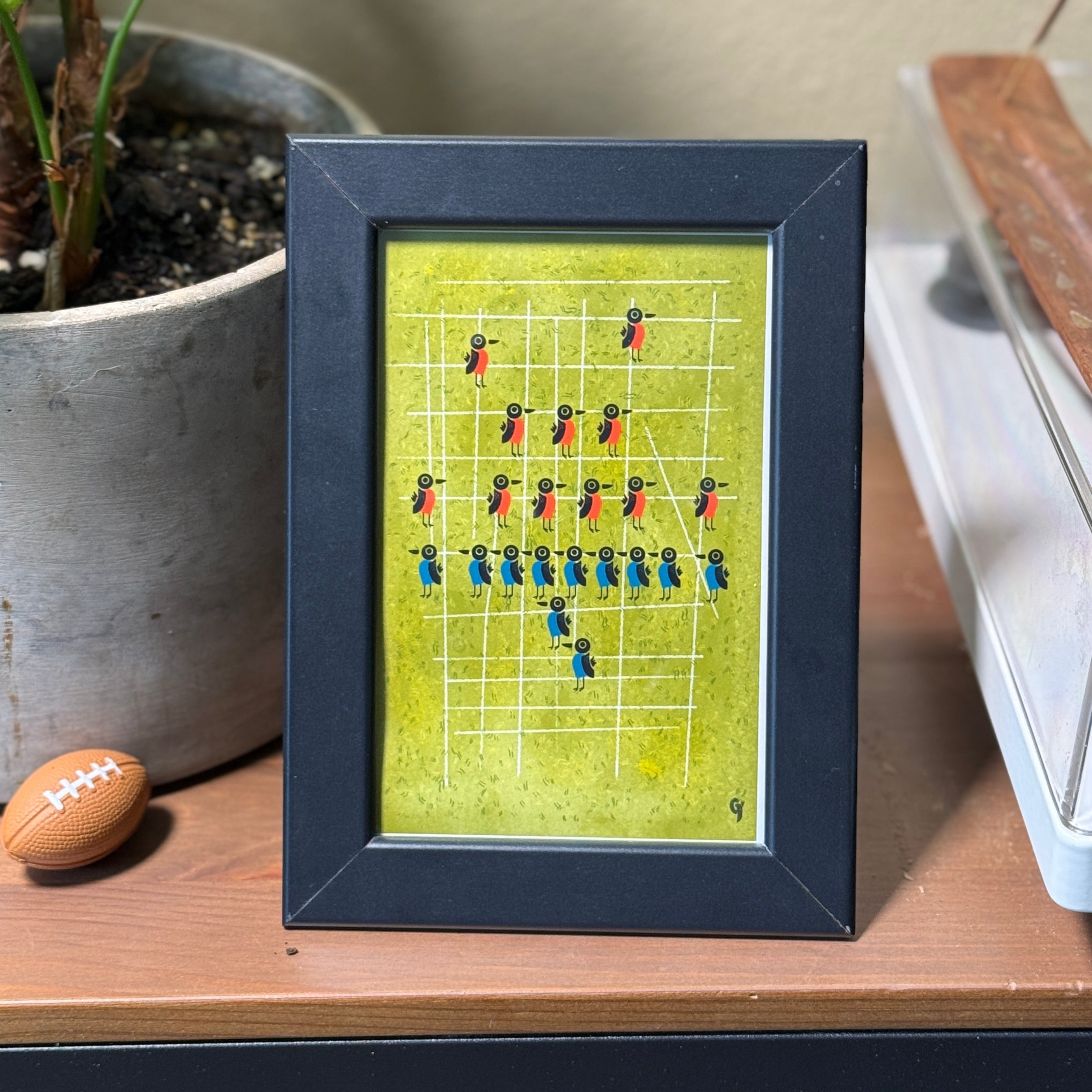 Framed artwork of a sports team lineup on a green background, placed on a wooden surface with a plant and other items in the background.