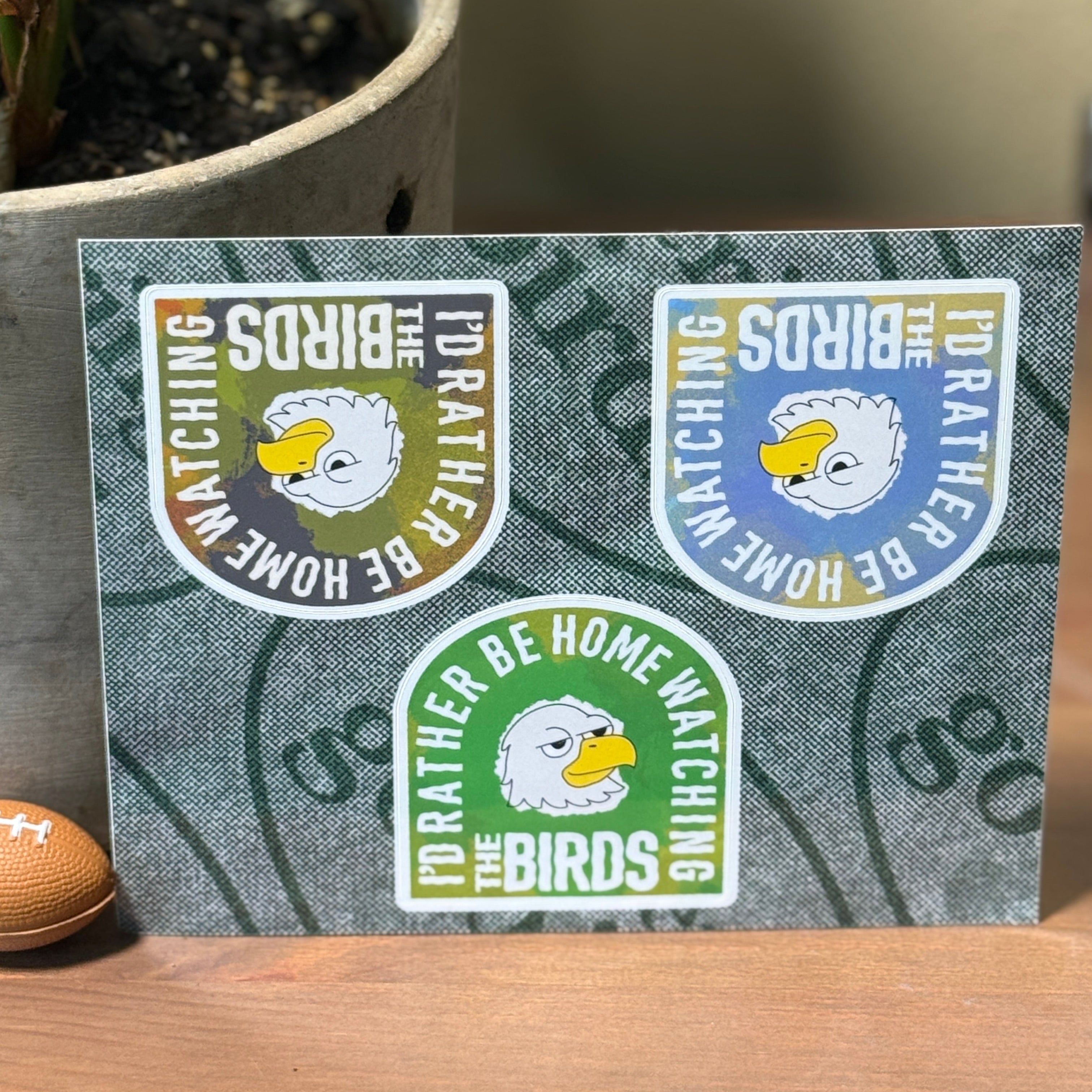 Three stickers with bird-themed designs on a textured card, placed on a wooden surface with a plant in the background.
