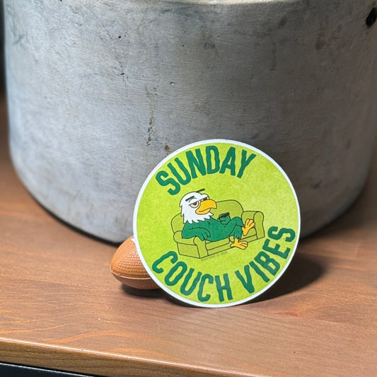 Round green sticker with 'Sunday Couch Vibes' text and an eagle graphic on a wooden surface next to a potted plant.