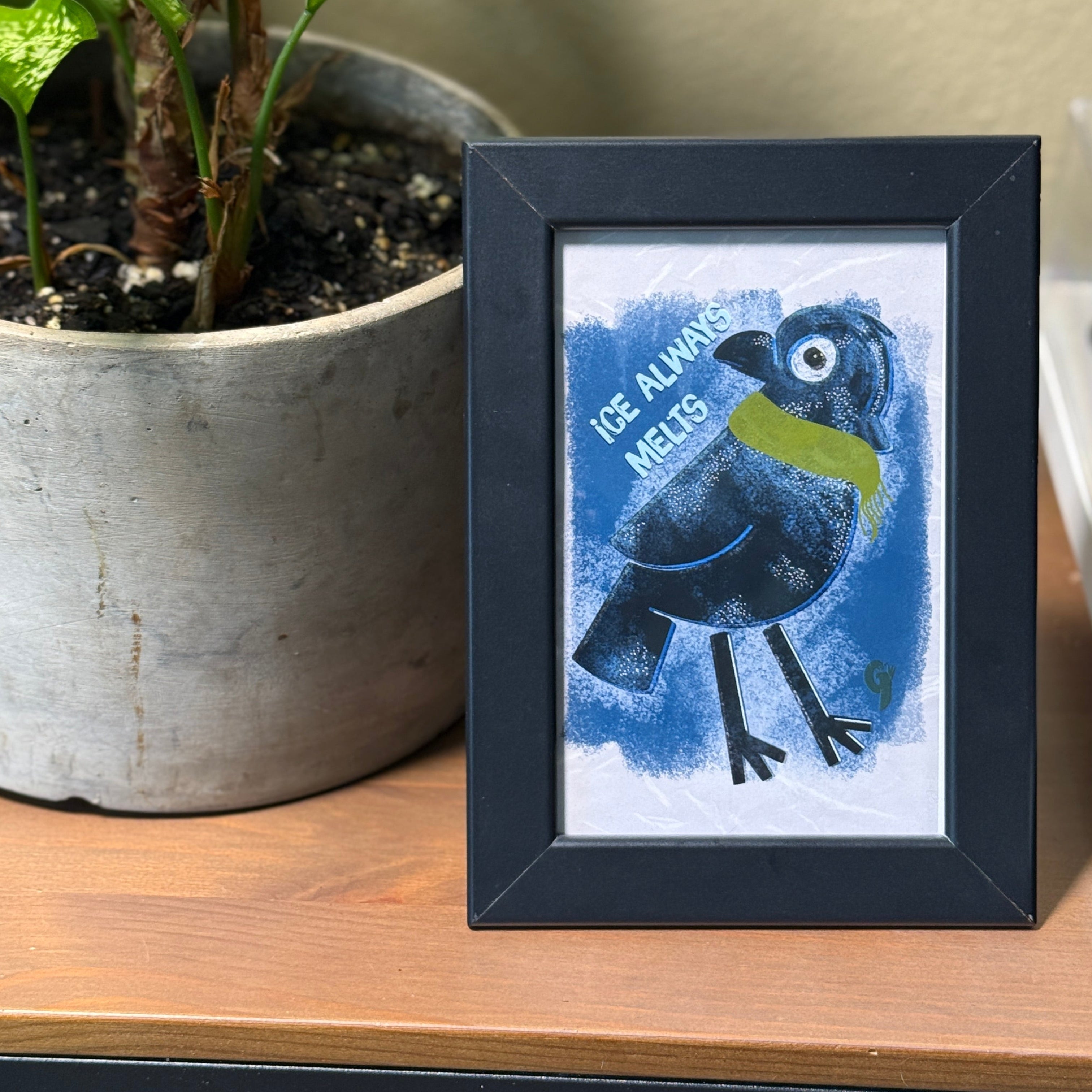 Framed artwork of a blue bird with text on a wooden surface next to a plant.