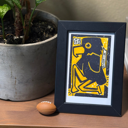 Framed artwork with a yellow and black design on a wooden surface next to a plant.
