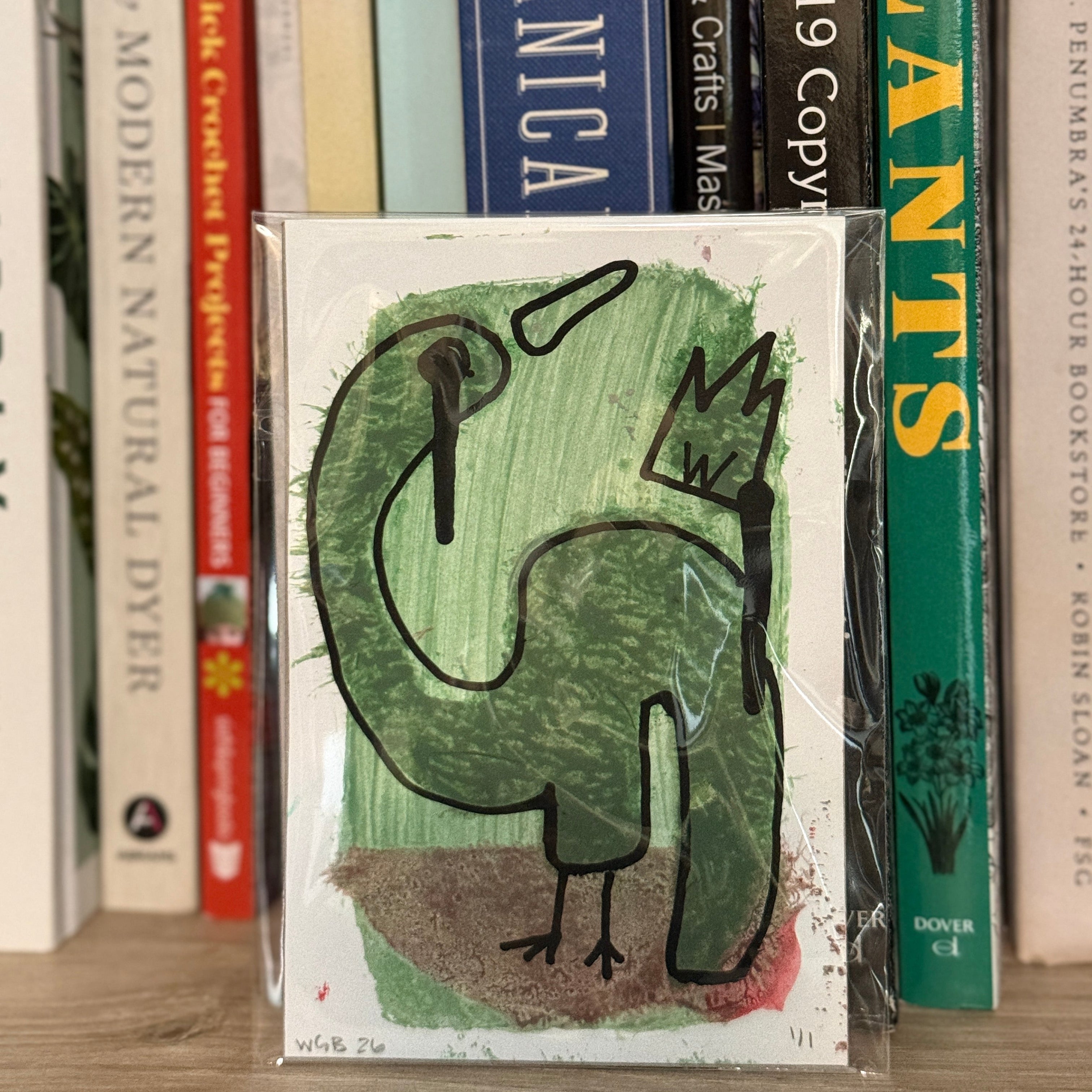 Card with a green dinosaur illustration on a bookshelf with various books.