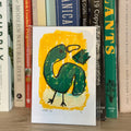 Card with a green bird design on a yellow background, placed among books on a shelf.