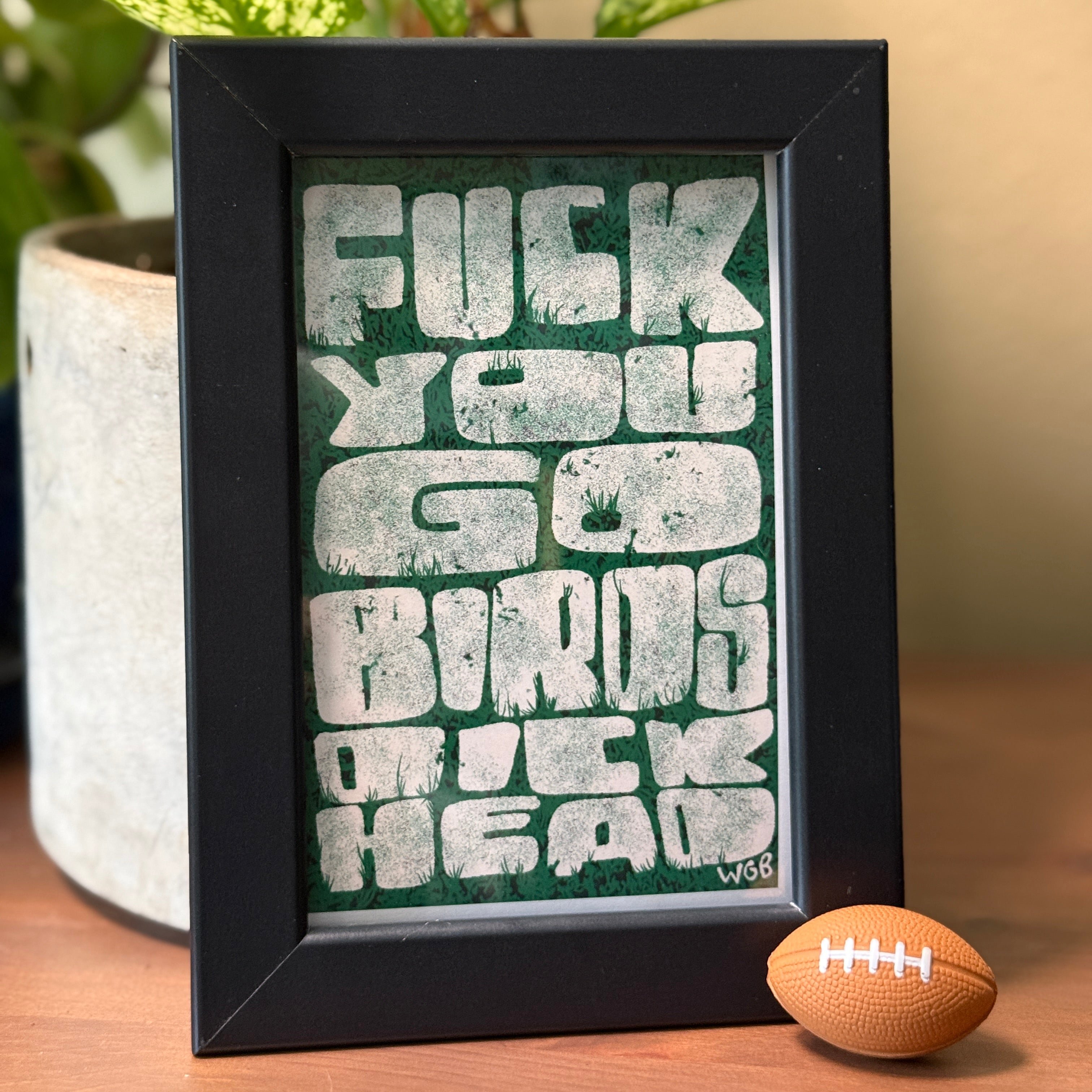 Framed artwork with offensive language on a table next to a football and plant.